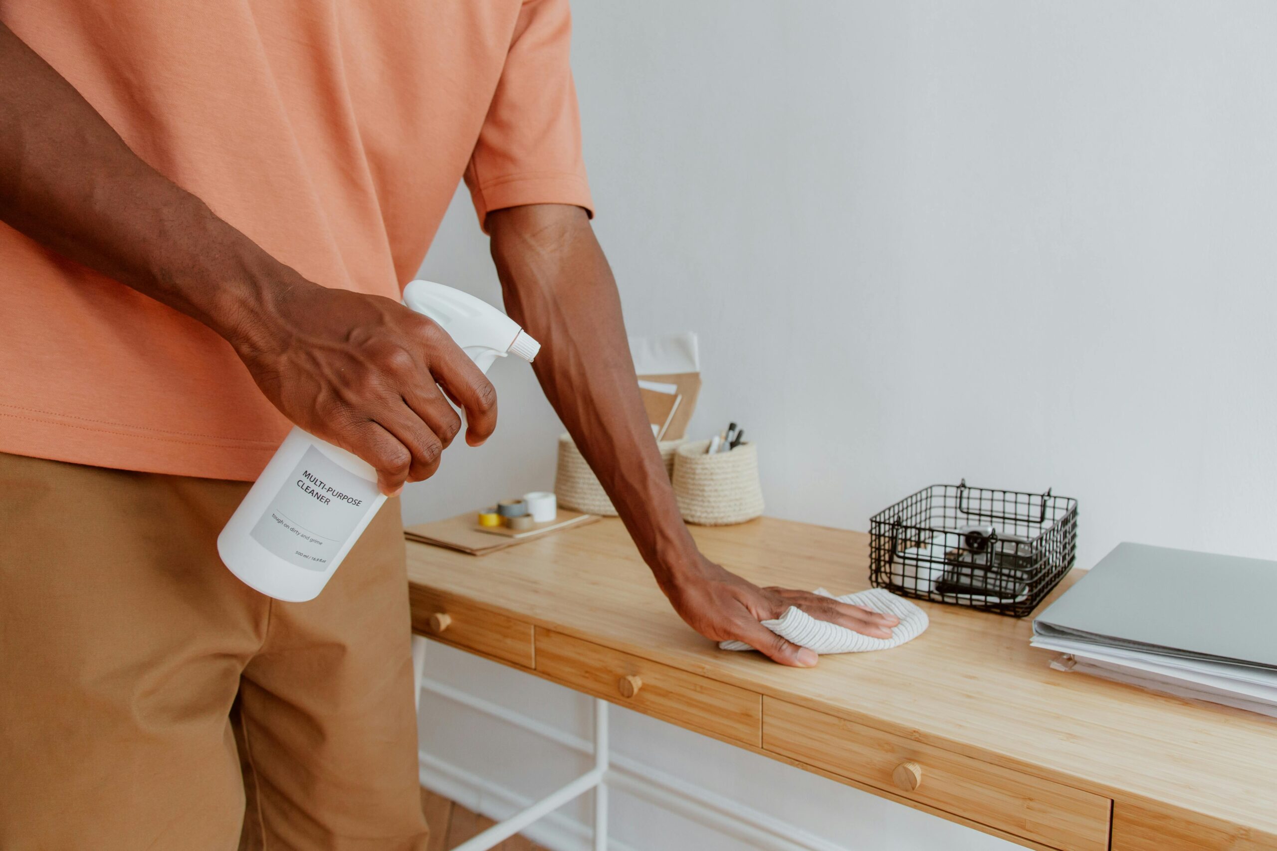 A person cleaning a wooden desk with a spray bottle and cloth, showcasing tidiness.