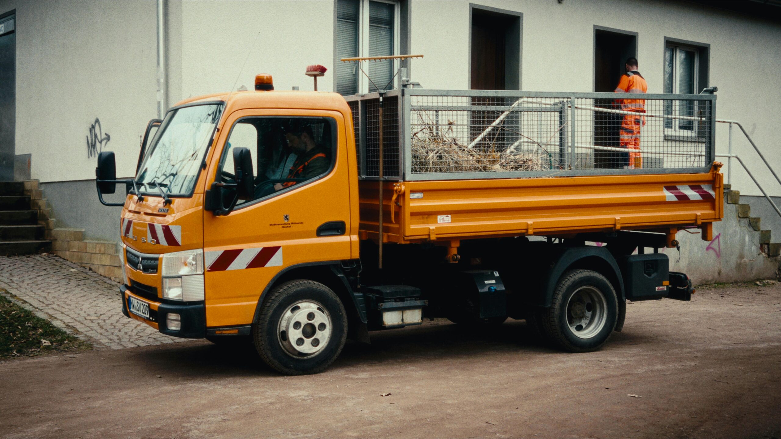 Orange utility truck parked near a building with workers wearing high-visibility clothing.